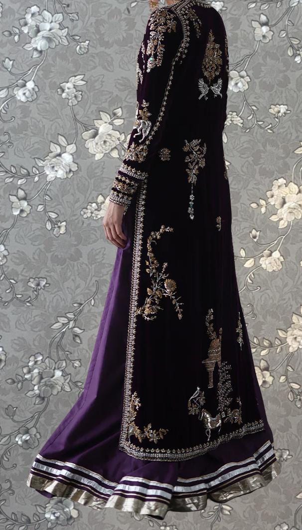 A person wearing a long, purple dress with intricate gold embroidery and a matching jacket stands against a gray floral background.