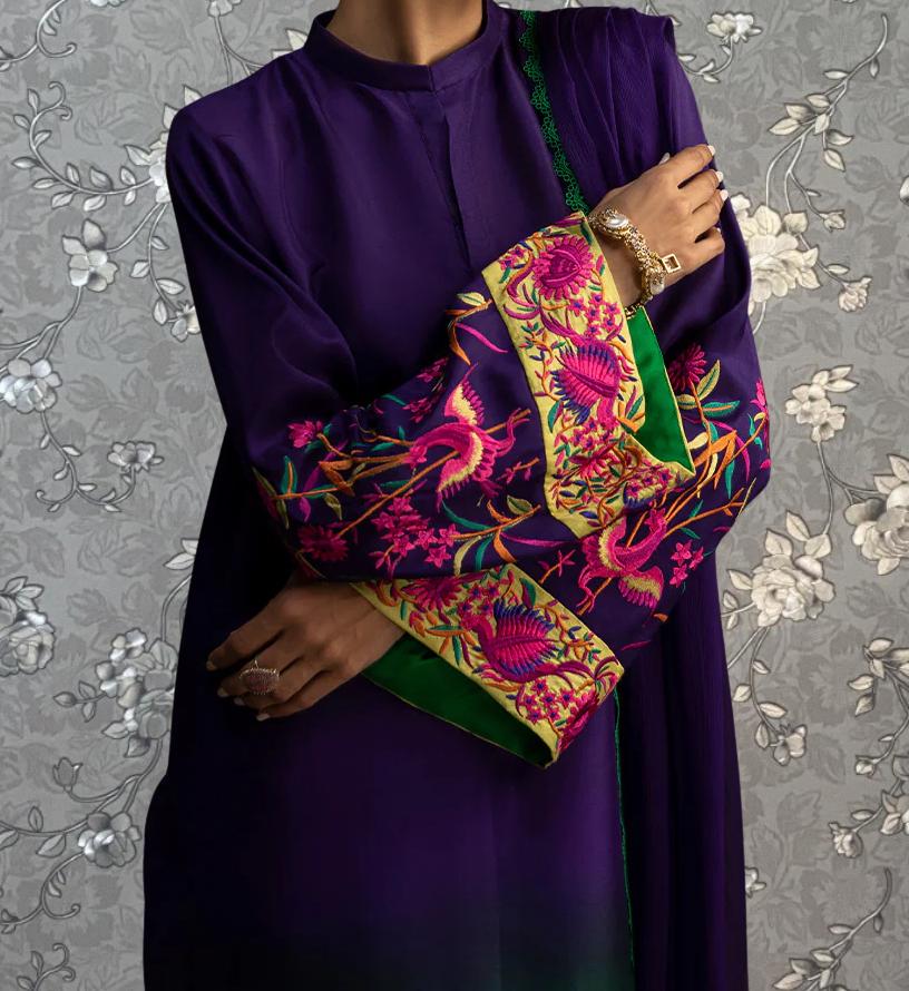 A person wearing a purple dress with a floral pattern and a green and yellow trim, posing with their arms crossed.