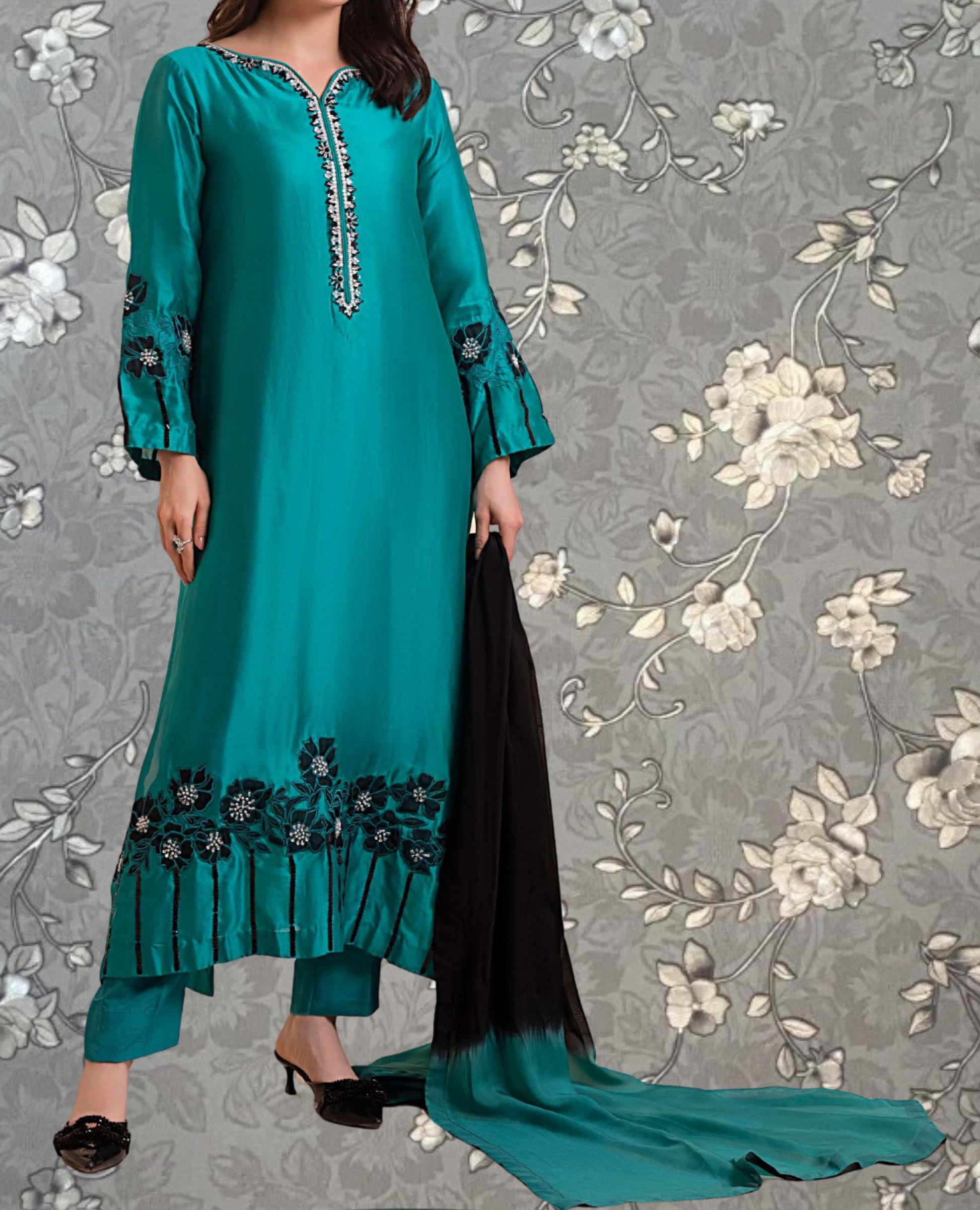 A woman wearing a long, turquoise dress with floral embroidery and holding a black shawl.