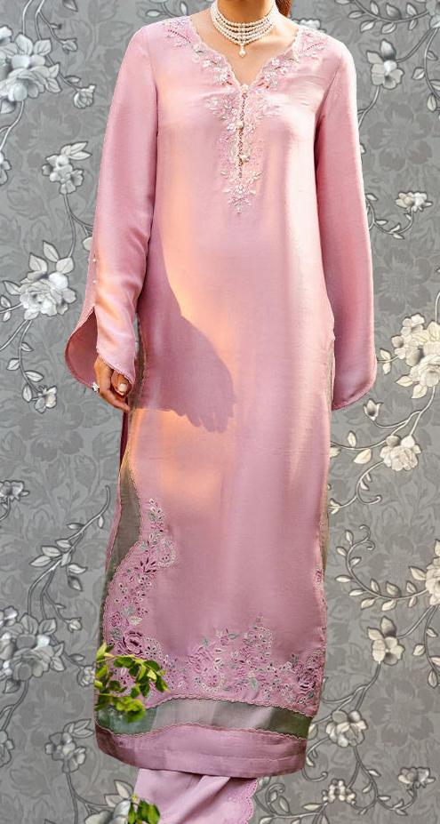 A woman wearing a long, pink dress with intricate embroidery and floral patterns, standing against a gray floral background.
