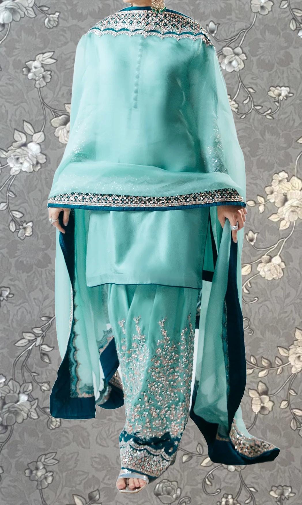 A turquoise and blue traditional Indian outfit with intricate embroidery and patterns, displayed against a gray floral background.