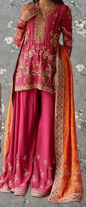 A woman wearing a vibrant pink and orange traditional Indian outfit, including a long skirt and a matching shawl, stands against a gray floral background.
