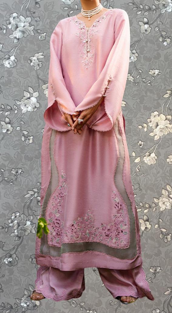 A woman wearing a long pink dress with intricate embroidery stands against a gray floral background.