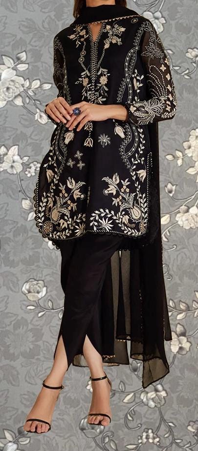 A woman wearing a black and gold embroidered dress with a floral pattern, standing against a gray floral background.