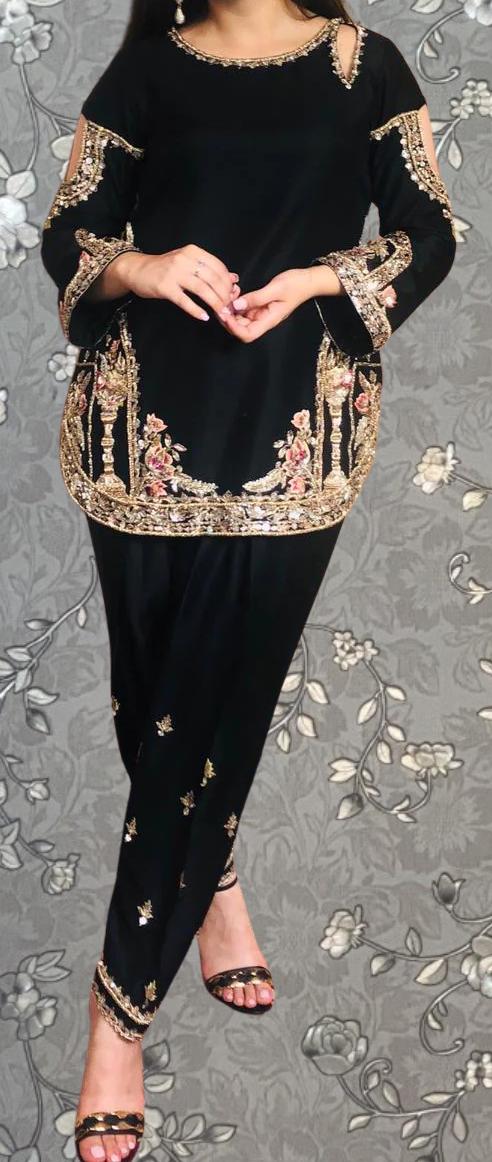 A woman wearing a black dress with gold embroidery and floral patterns, standing against a gray floral background.