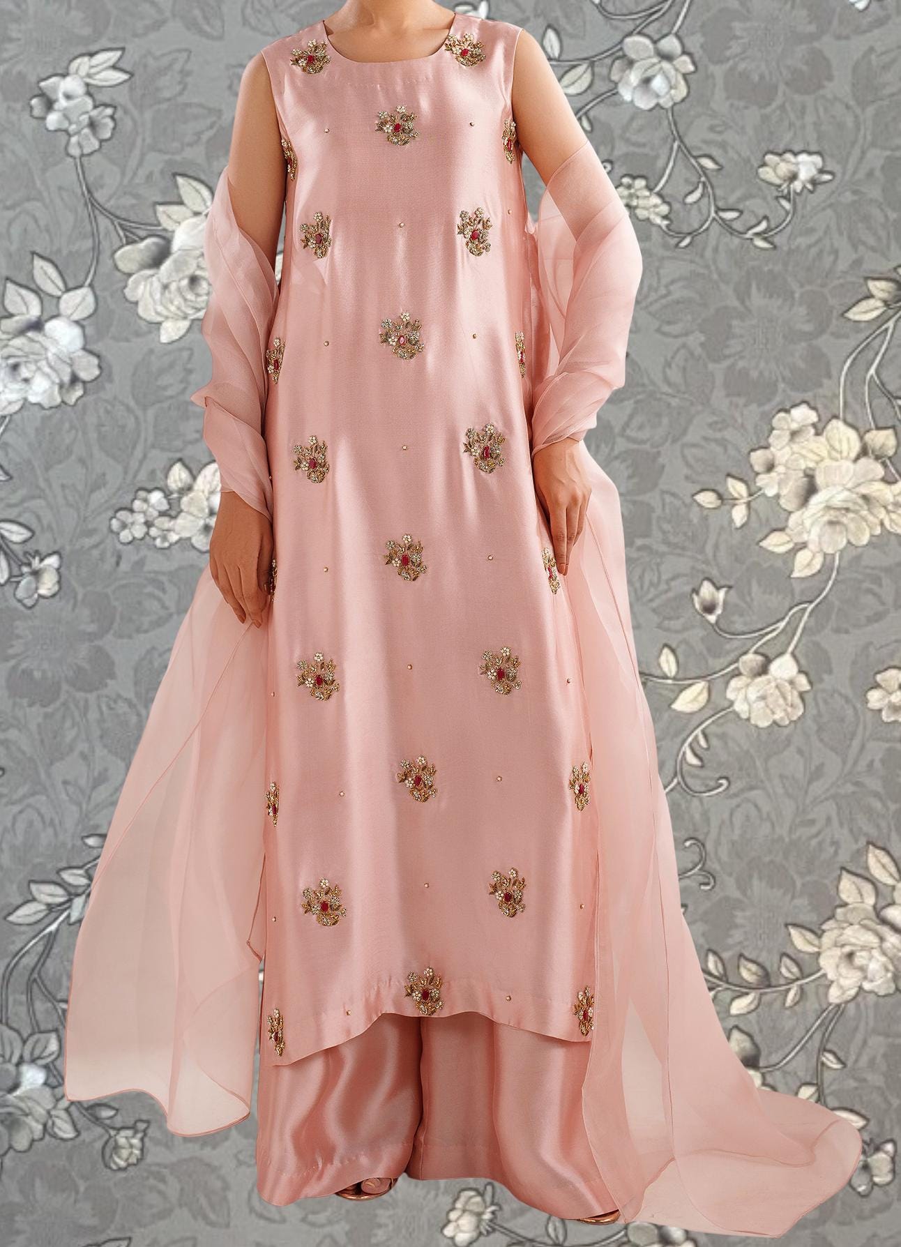 A woman wearing a pink dress with floral embroidery and a matching pink shawl stands against a gray floral background.