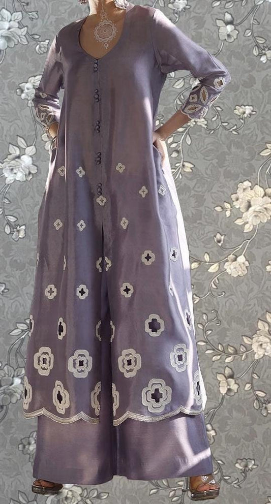 A woman wearing a long, purple dress with white floral embroidery stands against a gray floral background.