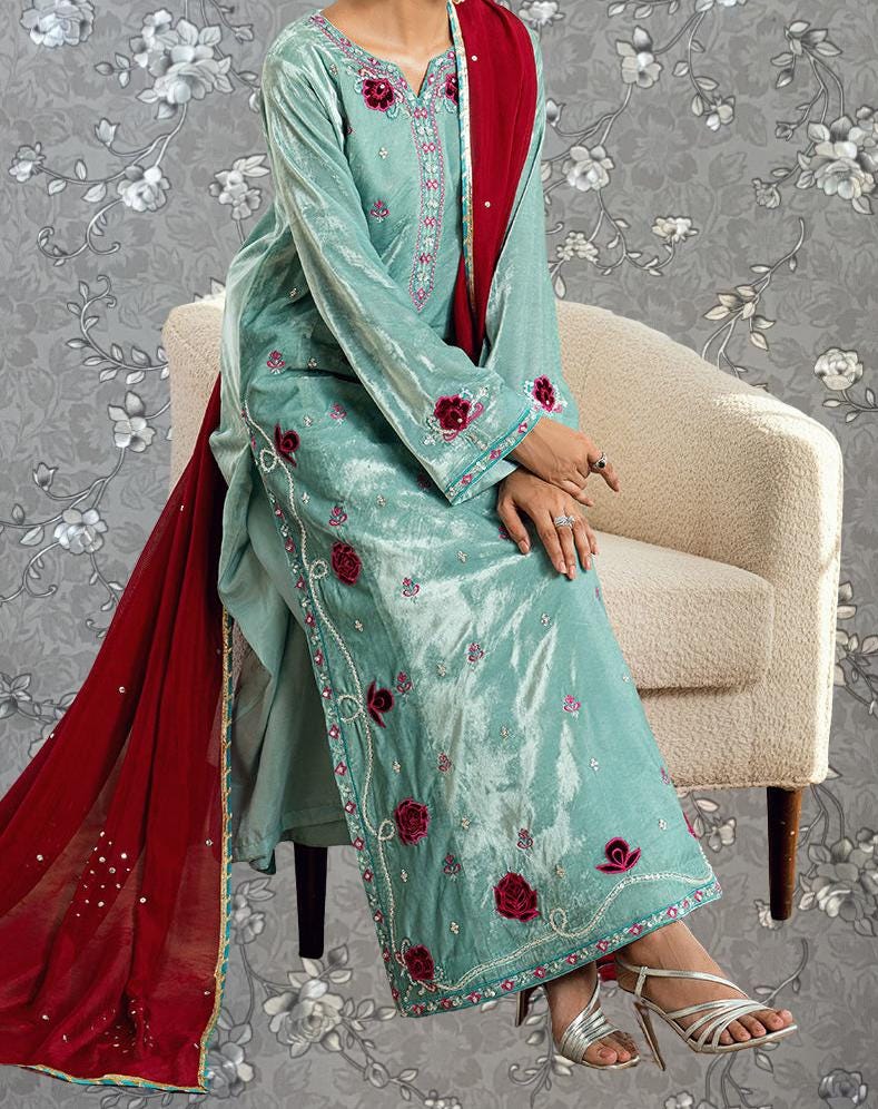 A woman wearing a long, light blue dress with red floral embroidery is sitting on a white chair against a gray floral background.