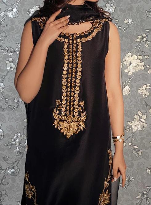 A woman wearing a black dress with gold embroidery stands against a gray floral background.