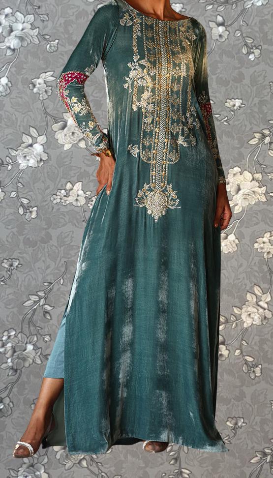 A woman wearing a long, green dress with intricate gold embroidery stands against a gray floral background.