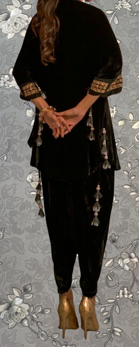 A person wearing a black velvet outfit with gold accents, including a jacket and pants, stands with their hands behind their back against a floral background.