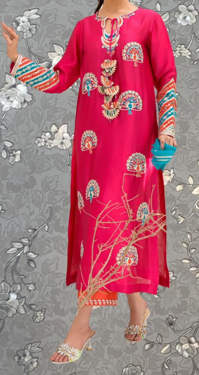 A person wearing a long pink dress with intricate embroidery and patterns, standing against a gray floral background.