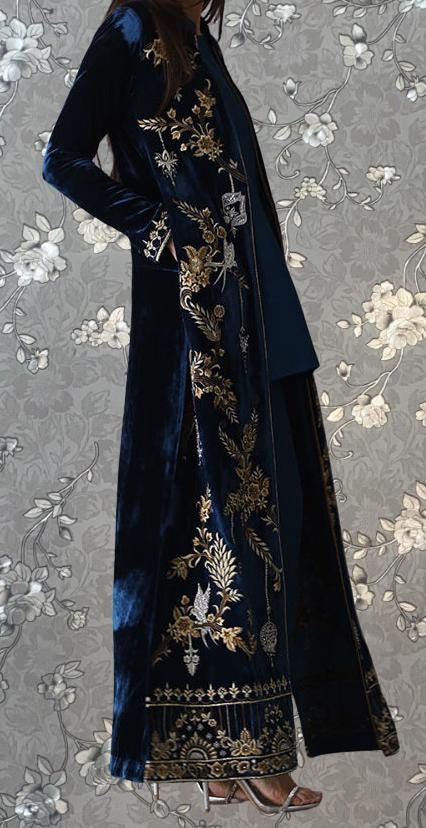 A woman wearing a long, navy blue velvet coat with gold embroidered patterns stands against a gray floral background.
