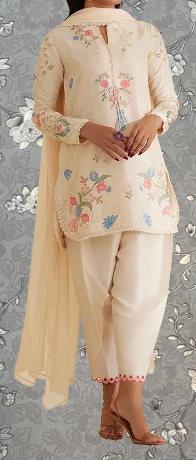 A woman wearing a cream-colored dress with floral embroidery stands against a gray floral background.