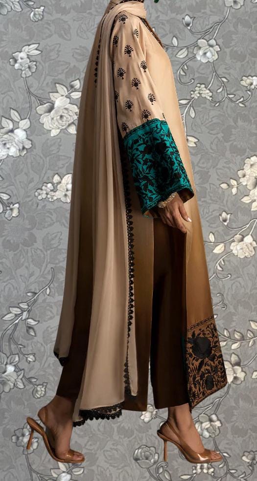 A person wearing a long, beige dress with a floral pattern and teal accents, standing against a gray floral background.