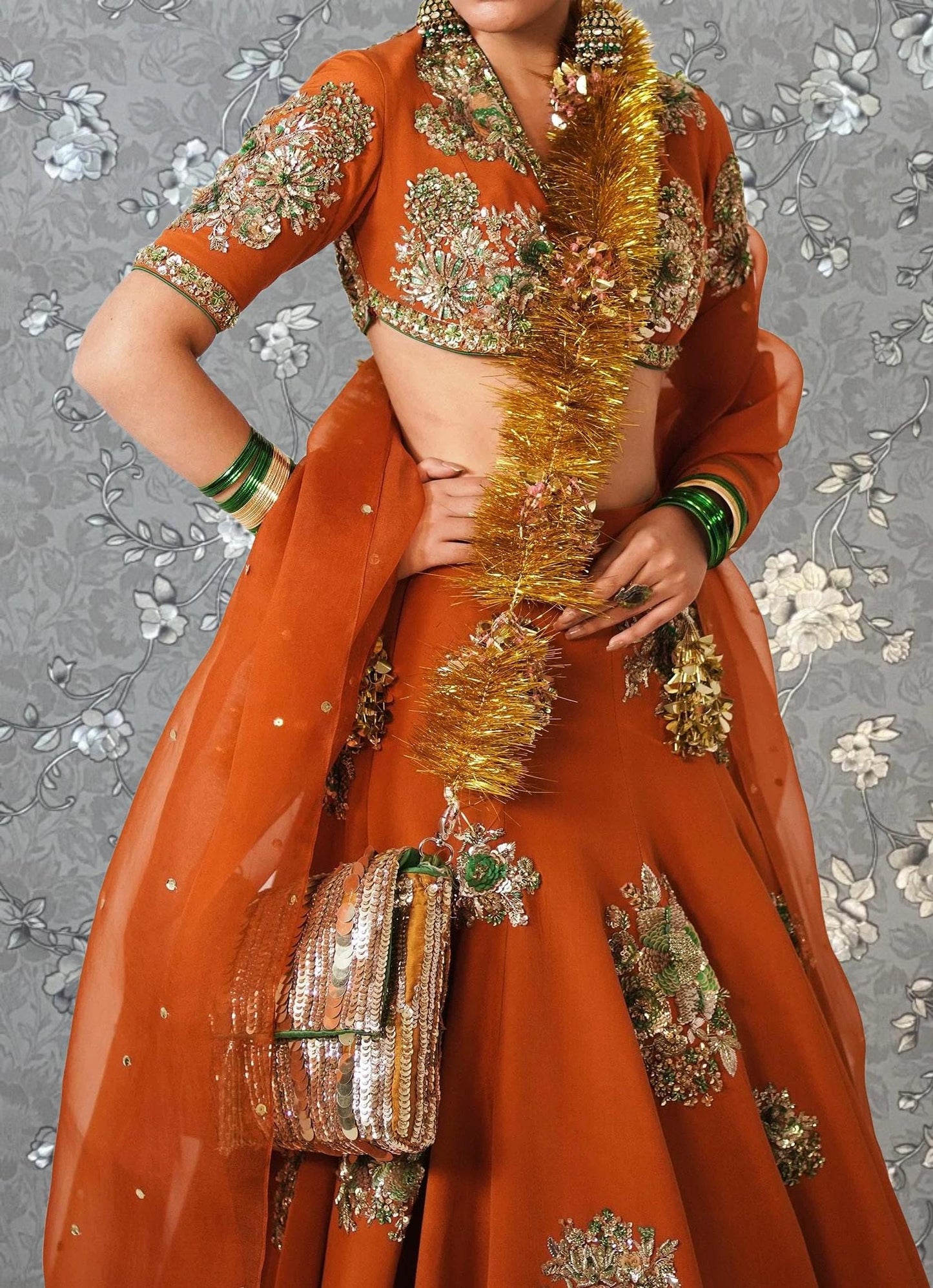 A woman wearing an ornate, brightly colored orange dress with gold and green embroidery, posing against a gray floral background.