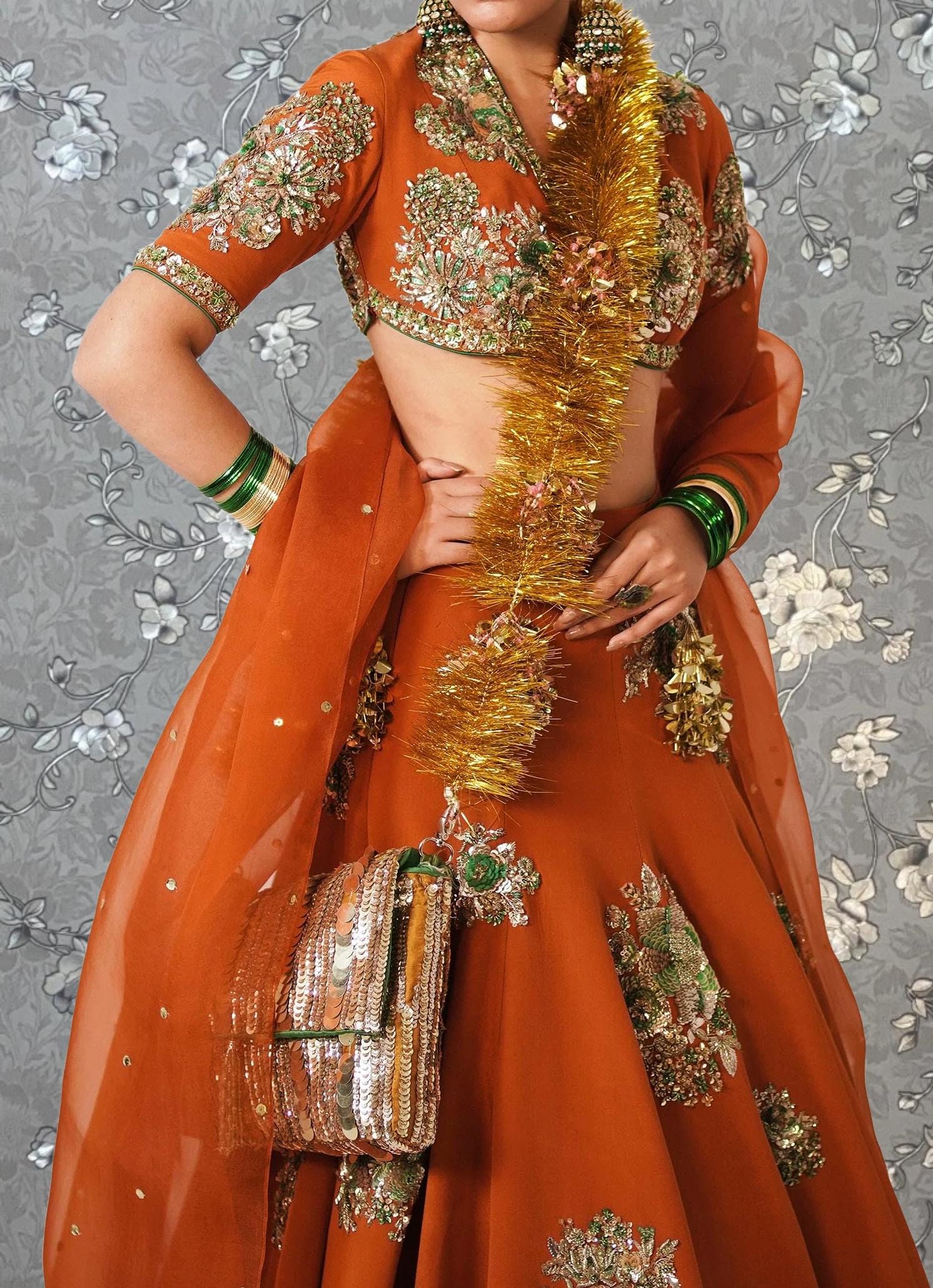A woman wearing an ornate, brightly colored orange dress with gold and green embroidery, posing against a gray floral background.