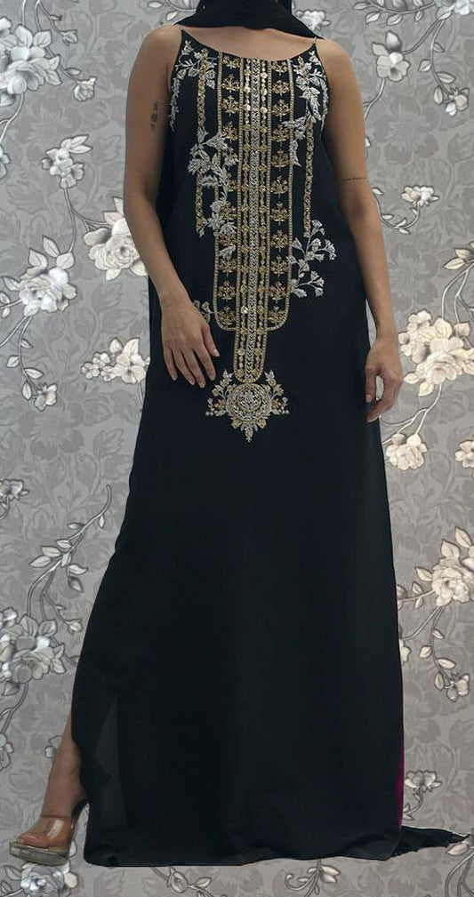 A woman wearing a long black dress with gold embroidery stands against a floral background.