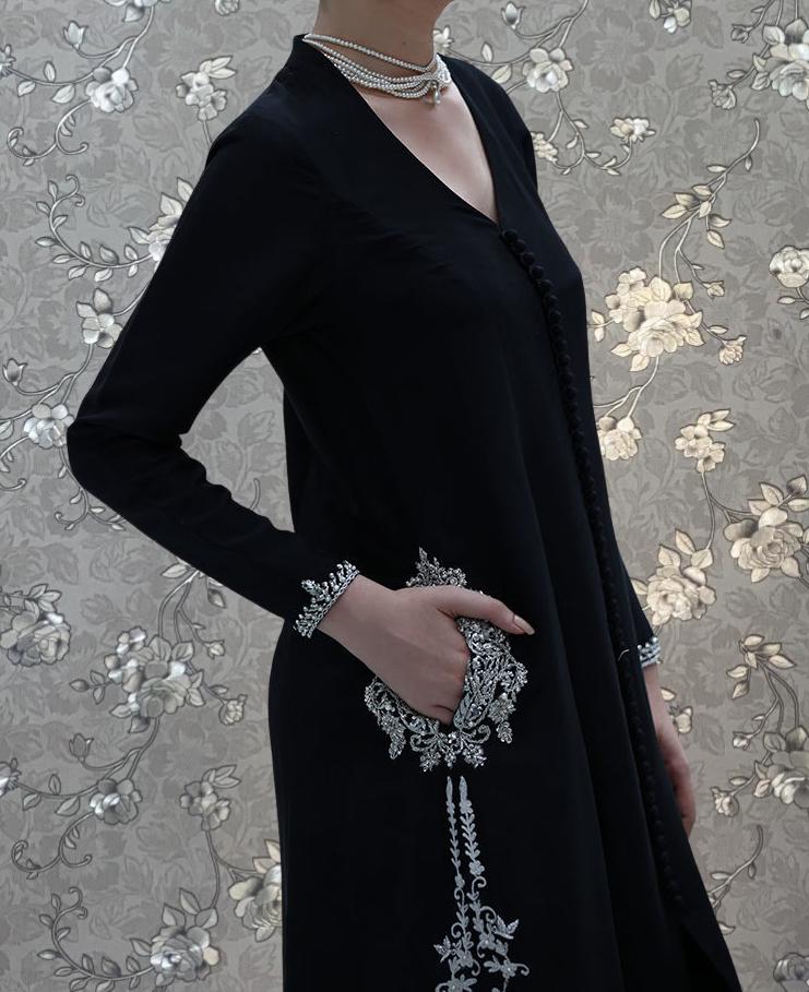 A woman wearing a black dress with intricate silver embroidery stands against a floral patterned background.