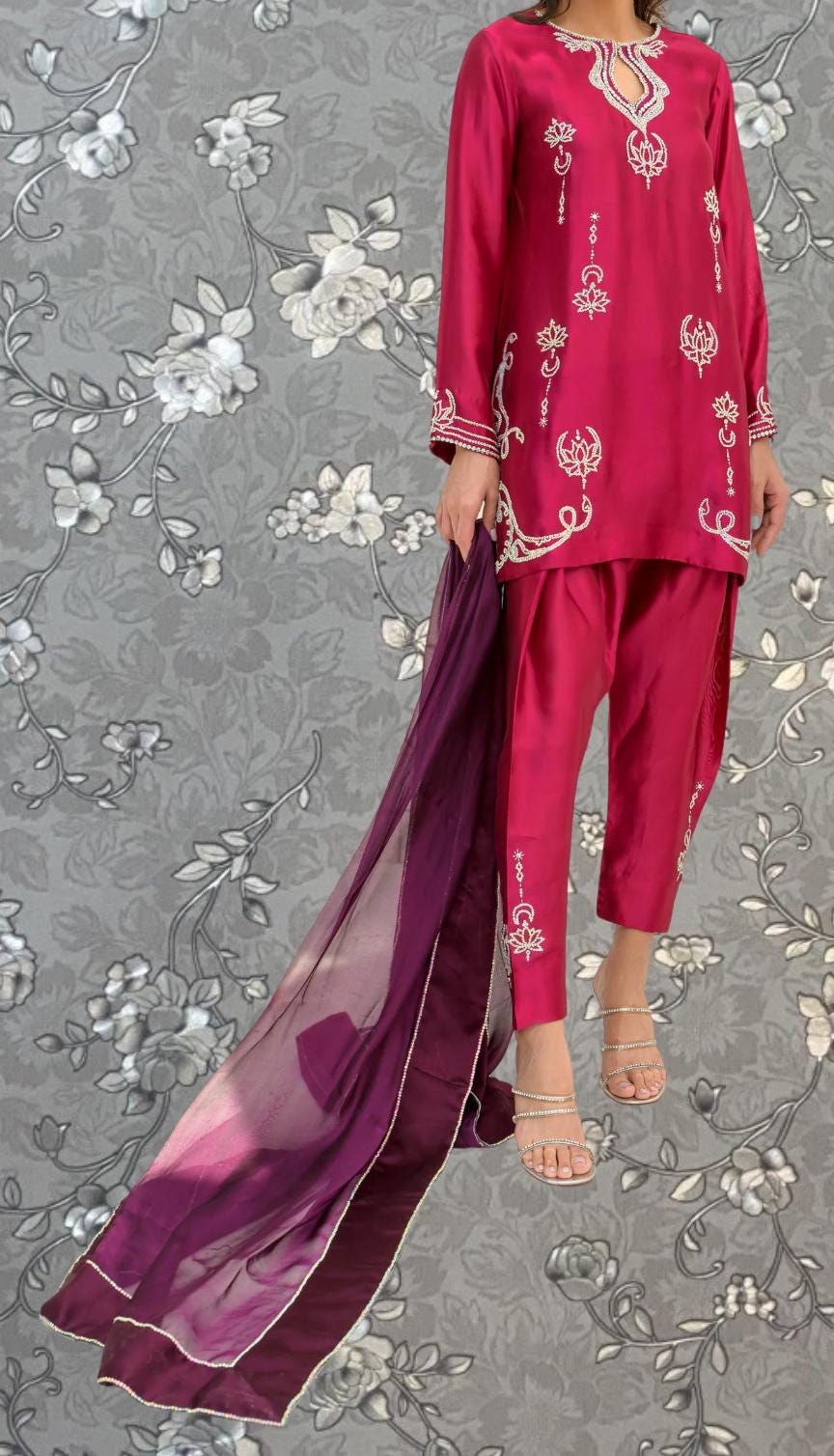 A person wearing a vibrant red outfit with intricate embroidery stands against a gray floral background.