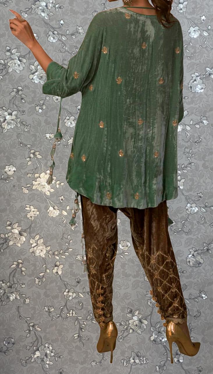 A person wearing a green velvet top with gold embroidered designs, brown velvet pants, and gold high heels is standing against a gray floral background.