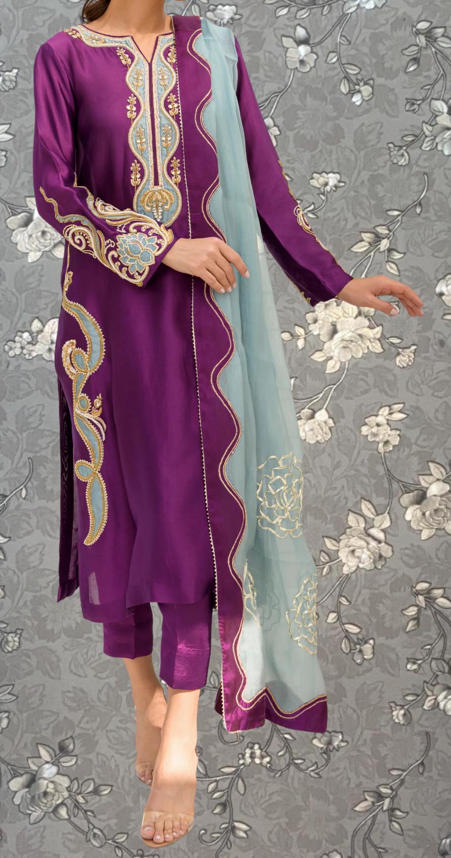 A person wearing a purple and blue dress with intricate embroidery and a matching shawl stands against a gray floral background.