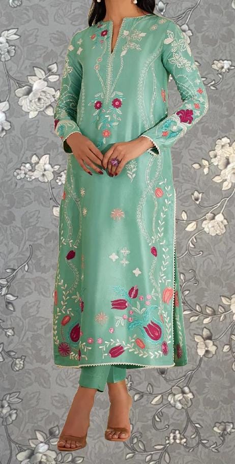 A woman wearing a long, mint green dress with intricate floral embroidery stands against a gray floral background.