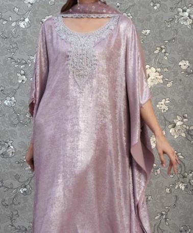 A woman wearing a long, flowing pink dress with intricate embroidery stands against a gray floral background.