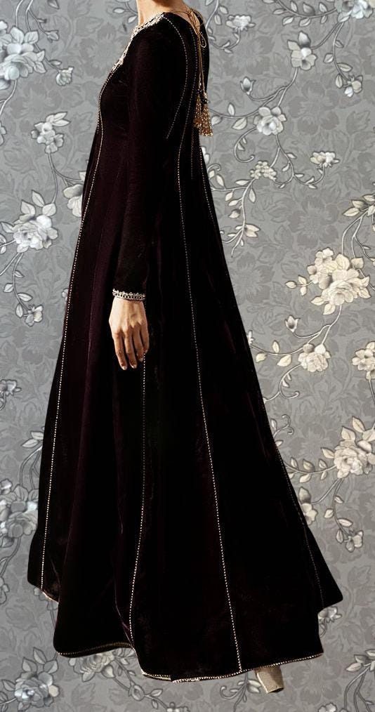 A person wearing a long, dark purple dress with a high collar and long sleeves stands against a gray floral background.