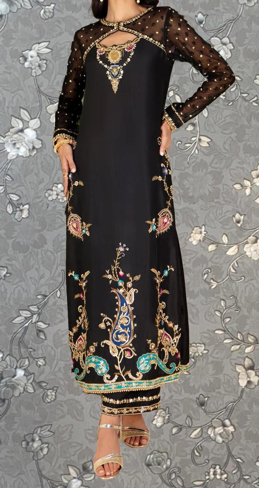 A woman wearing a long black dress with intricate gold embroidery and floral designs, standing against a gray floral background.