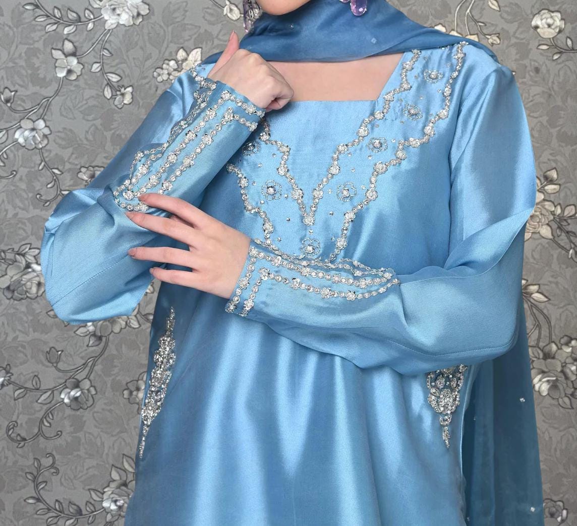 A person wearing a blue dress with intricate beading and embroidery details, posing with their hand on their chin.