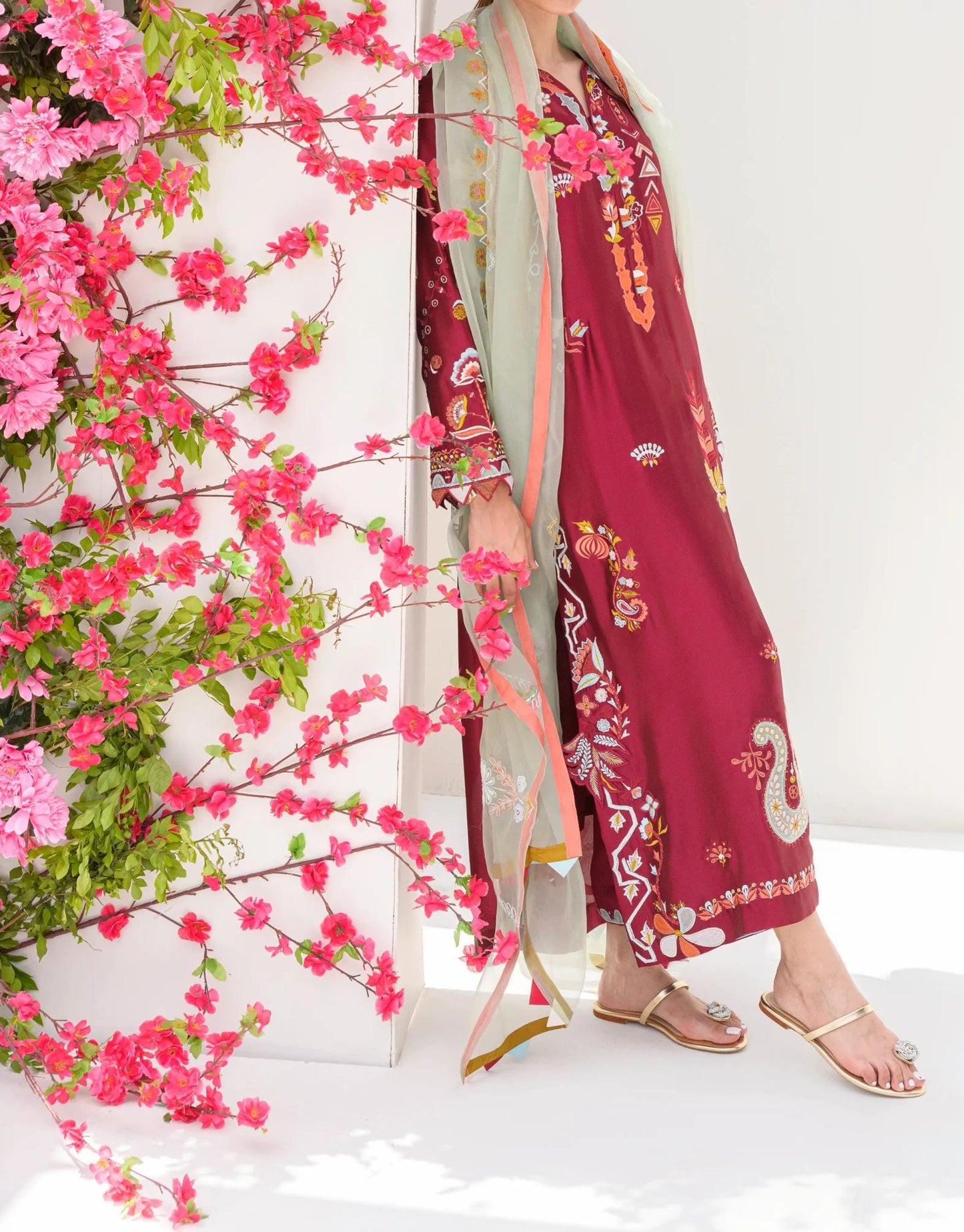A person wearing a long, burgundy dress with intricate floral embroidery stands in front of a white wall adorned with pink flowers.