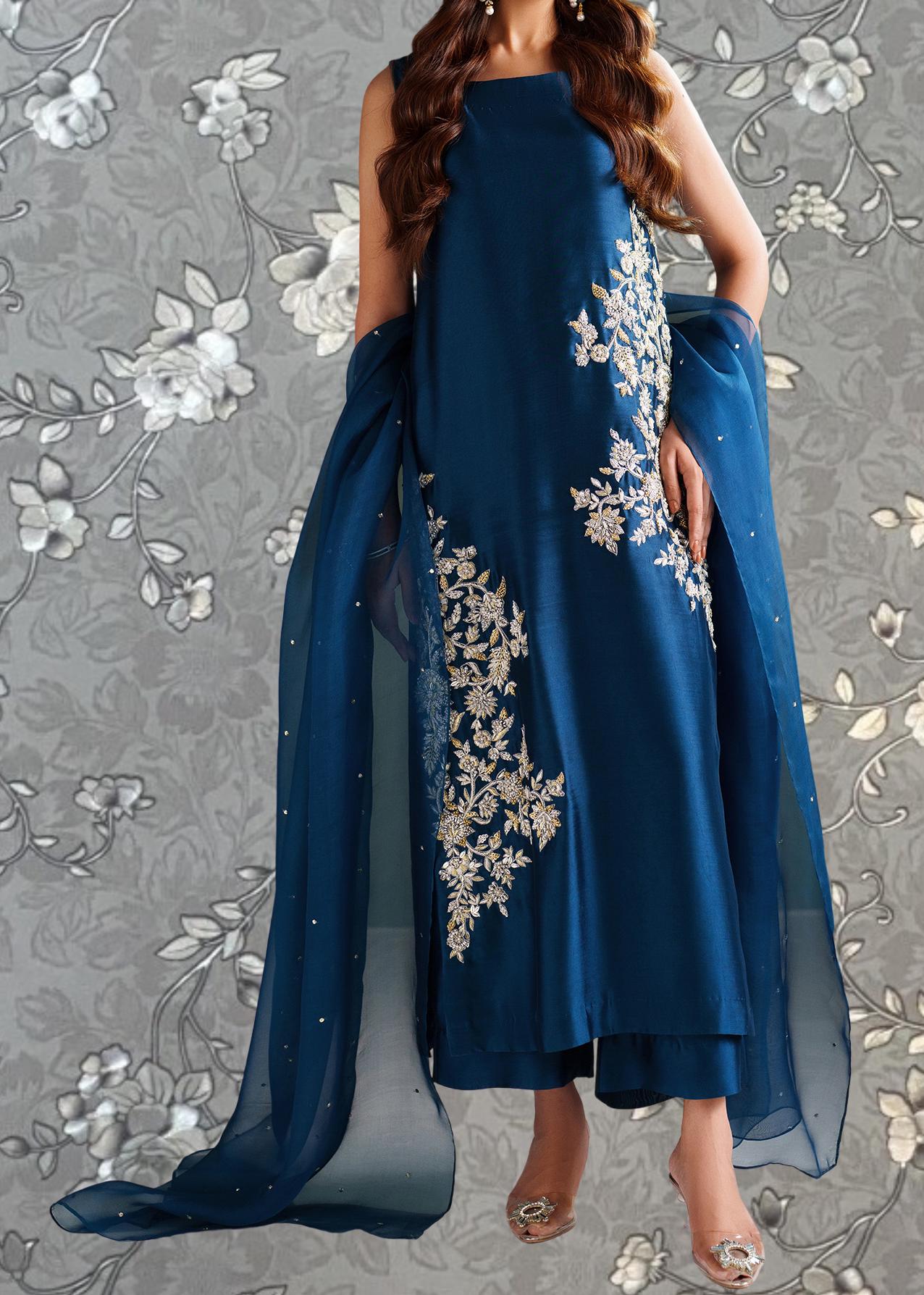 A woman wearing a long blue dress with intricate white floral embroidery stands against a gray floral background.