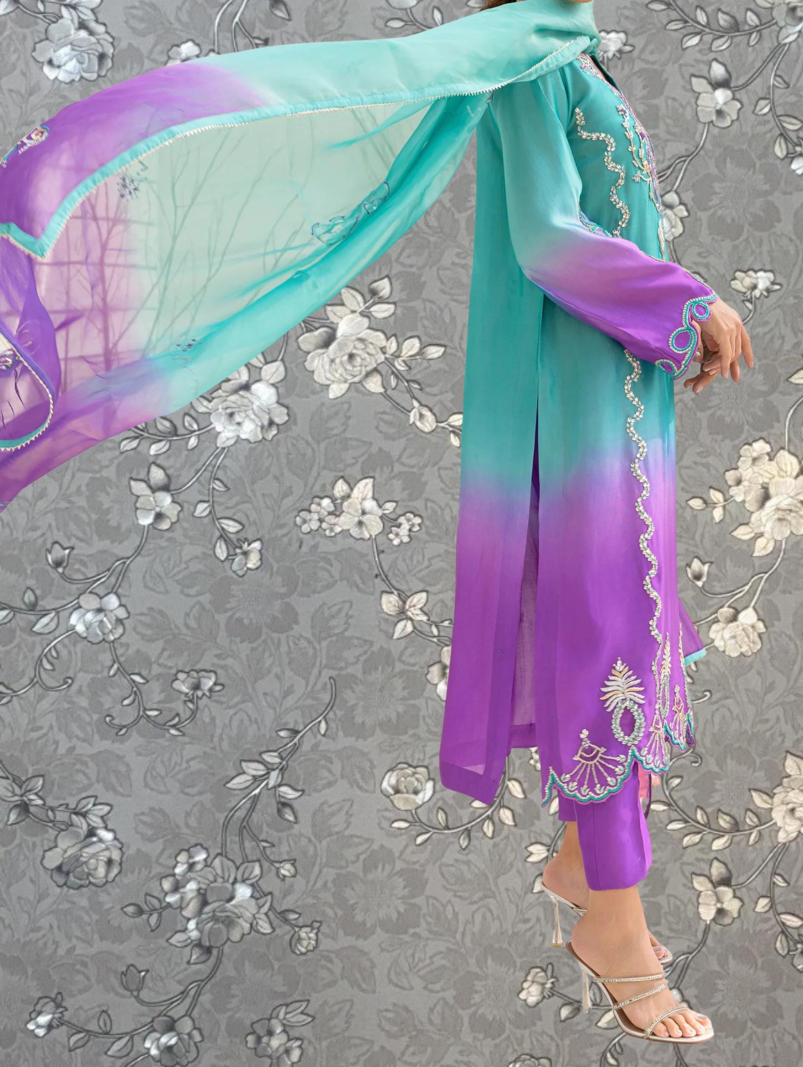 A person wearing a long, flowing dress with a purple and teal color scheme, standing against a gray floral background.