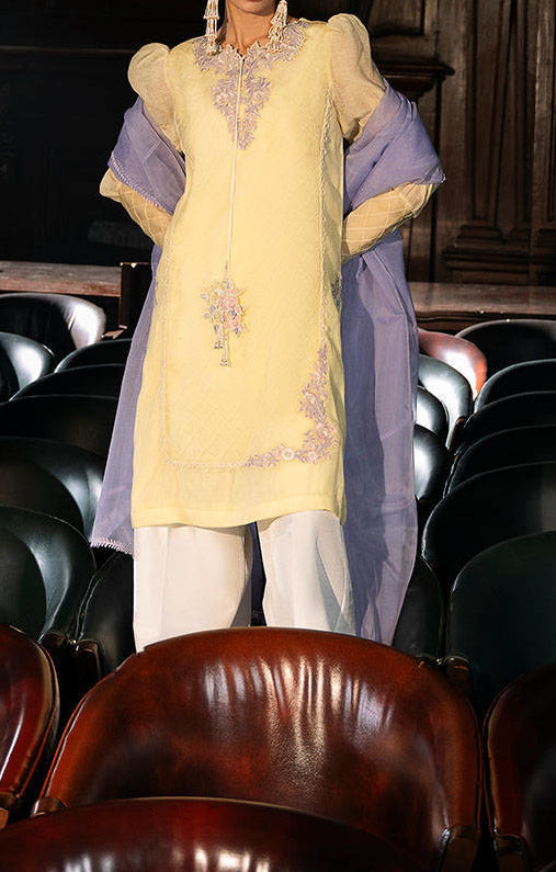 A person wearing a yellow dress with purple accents stands in a room with rows of chairs.