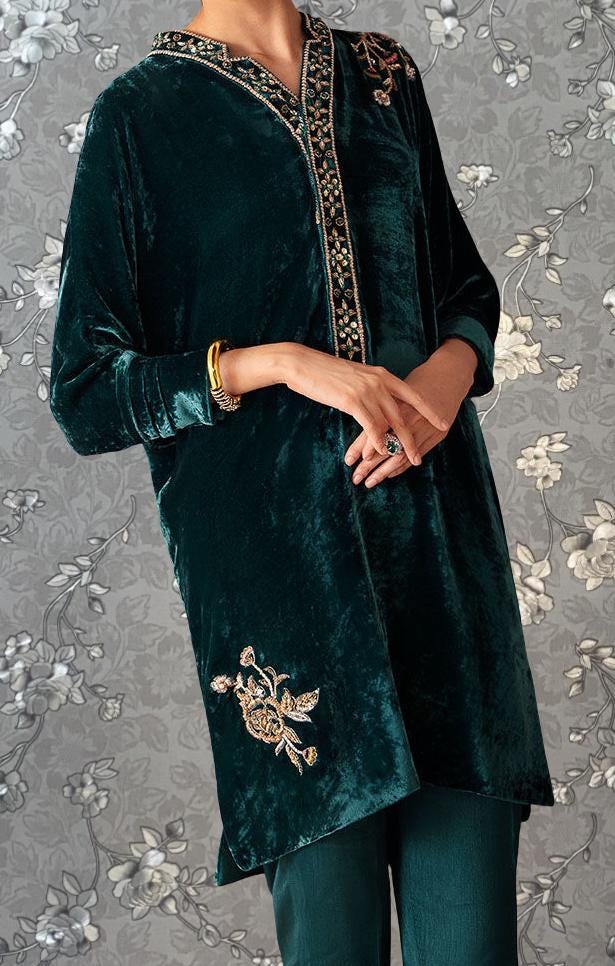 A person wearing a green velvet dress with gold embroidery stands against a gray floral background.