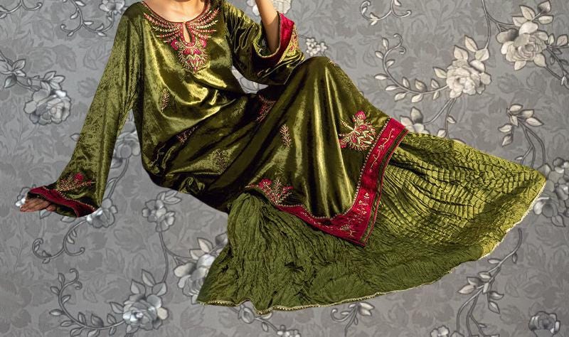 A woman wearing a long, green dress with red accents and floral embroidery, sitting against a gray floral background.