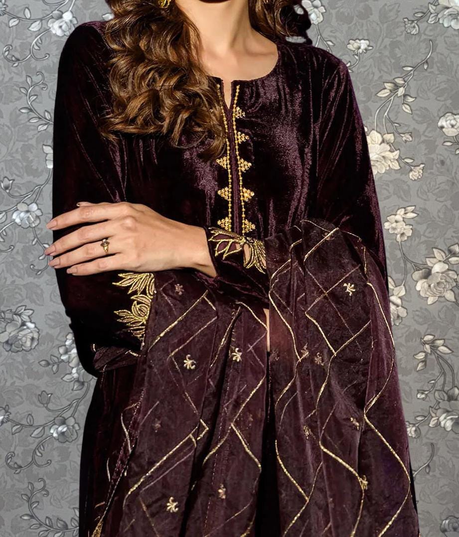 A woman wearing a long, burgundy velvet dress with gold embroidery stands against a gray floral background.