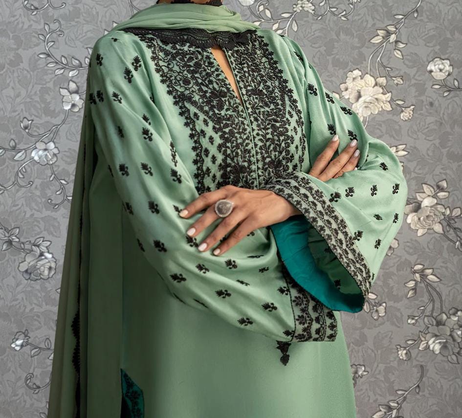 A person wearing a green and black patterned dress with a large floral design on the sleeves and collar, posing with their arms crossed.