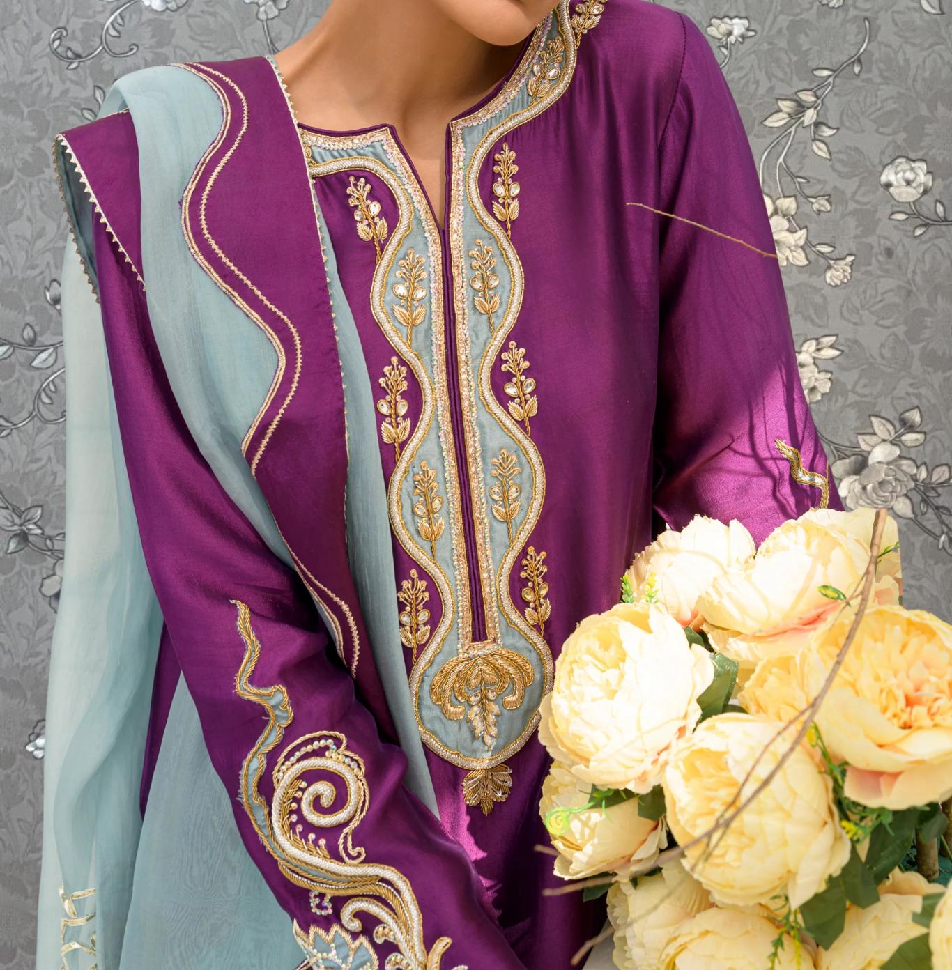 A woman wearing a purple and blue dress with intricate embroidery stands next to a bouquet of yellow flowers.