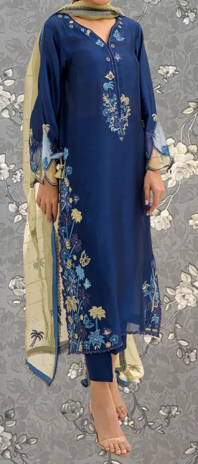 A woman wearing a blue dress with intricate embroidered designs, standing against a gray floral background.