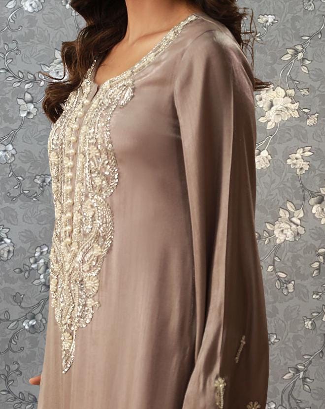 A woman wearing a long, beige dress with intricate embroidery and a high collar, standing against a floral patterned background.