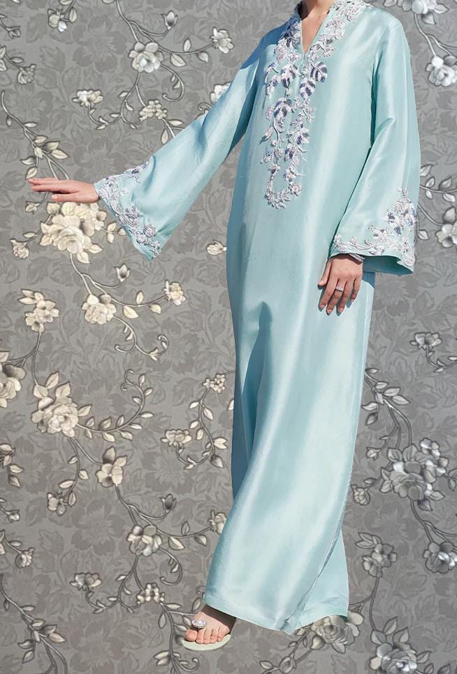 A person wearing a long, light blue dress with floral embroidery stands against a gray floral background.