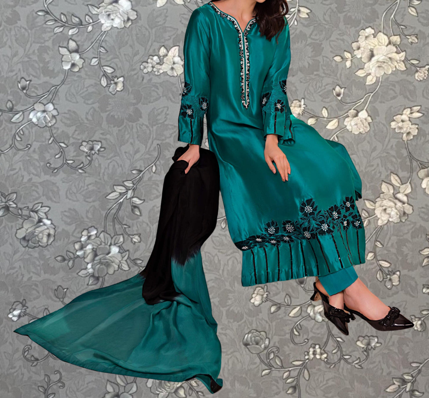 A woman wearing a long, green dress with floral embroidery and black pants is sitting on a gray floral background.