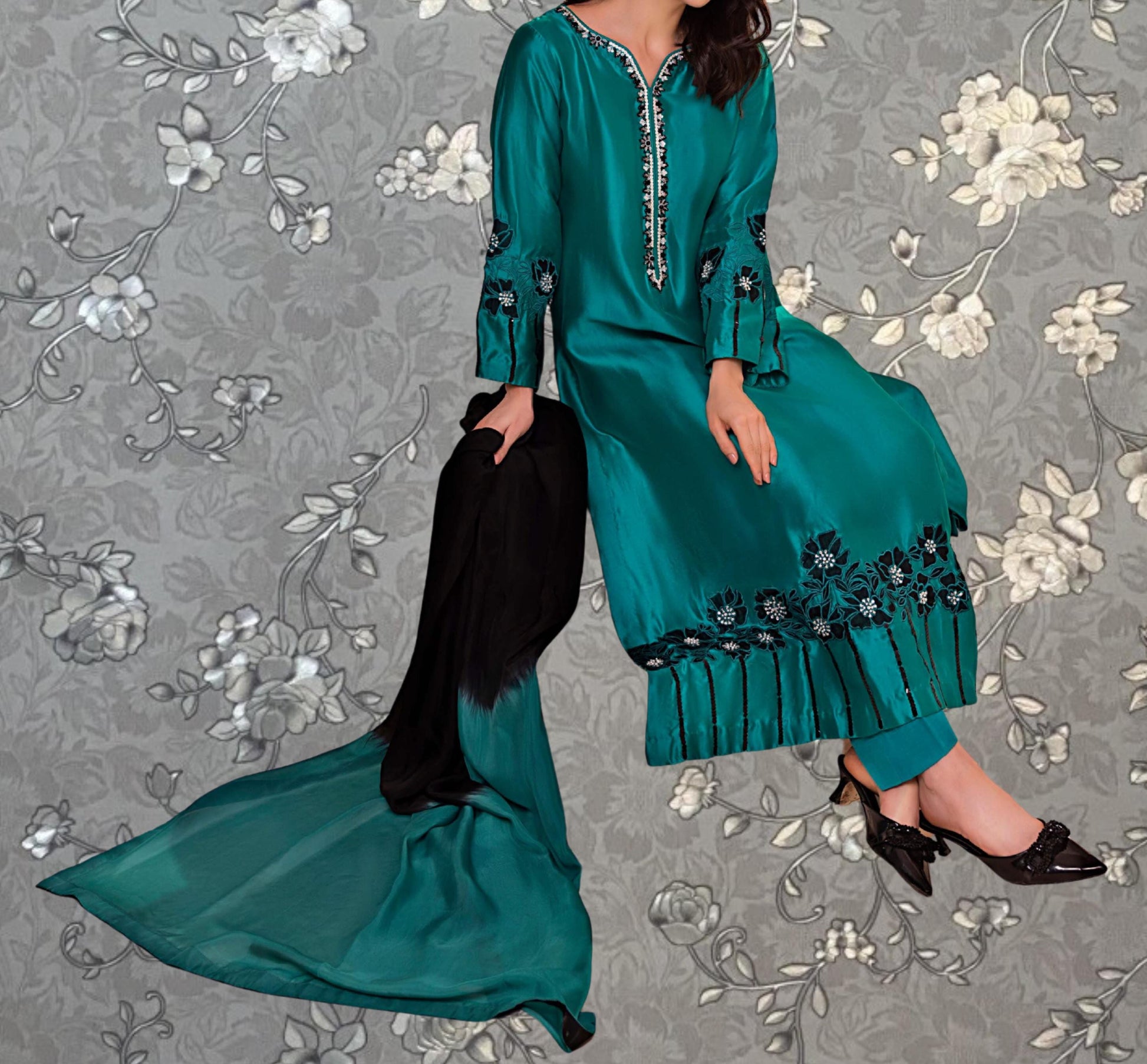 A woman wearing a long, green dress with floral embroidery and black pants is sitting on a gray floral background.