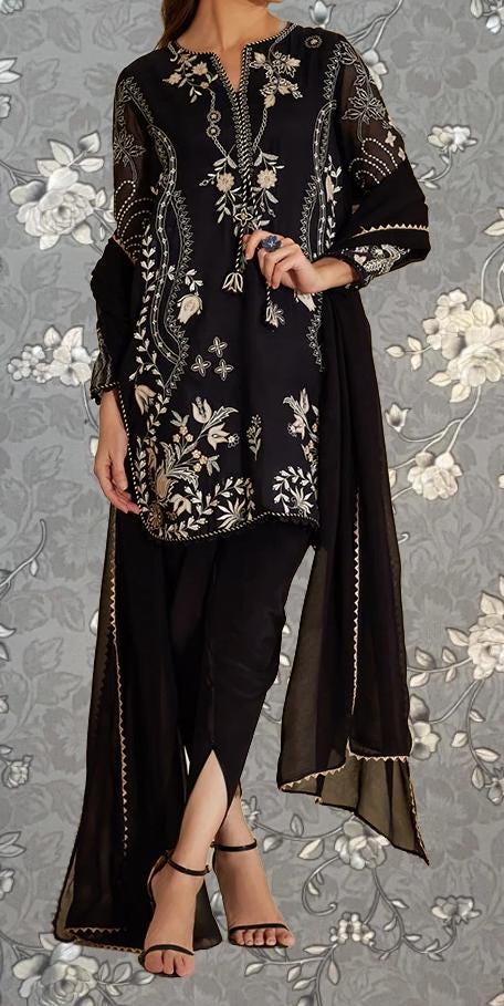 A woman wearing a black and gold patterned dress with a long black shawl draped over her shoulders, standing against a gray floral background.
