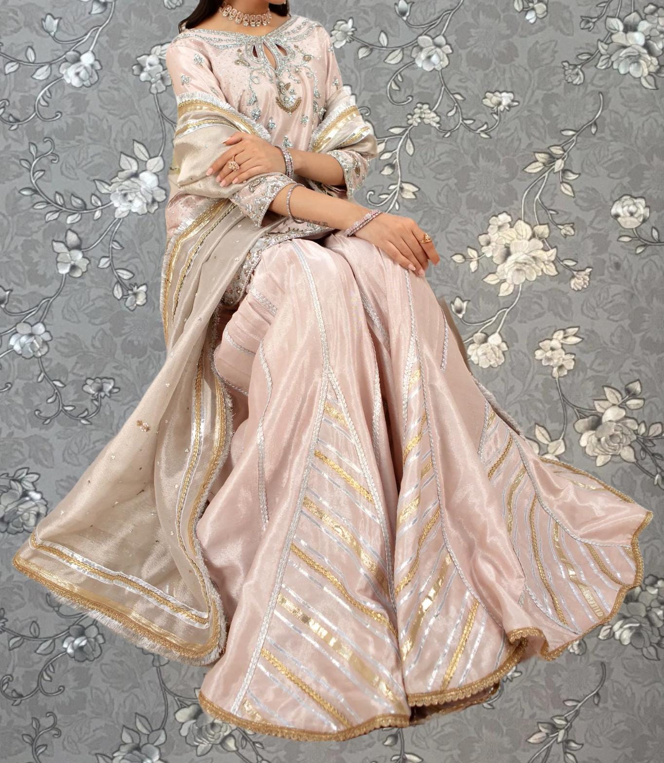 A woman wearing a pink and gold dress with intricate embroidery and a matching shawl is sitting against a gray floral background.
