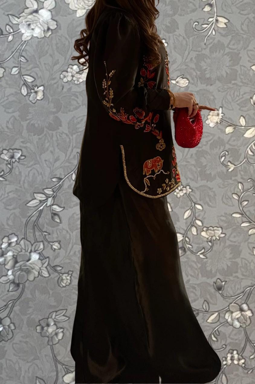 A woman wearing a long black dress with red embroidered designs, holding a red purse, standing against a gray floral background.