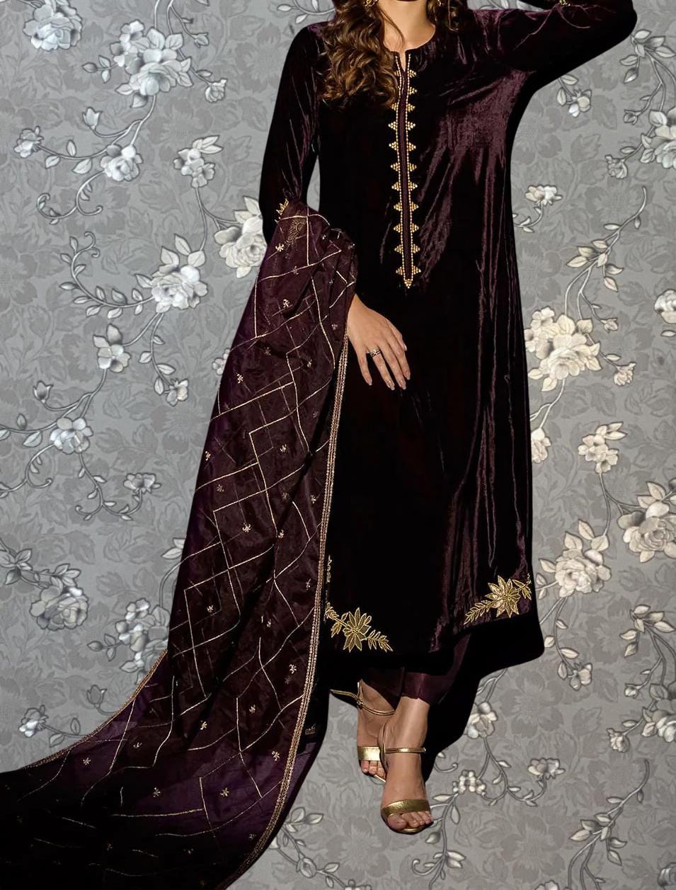 A woman wearing a long, burgundy velvet dress with gold embroidery stands against a gray floral background.