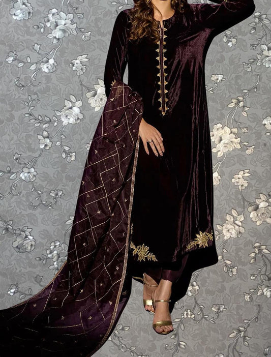 A woman wearing a long, burgundy velvet dress with gold embroidery stands against a gray floral background.